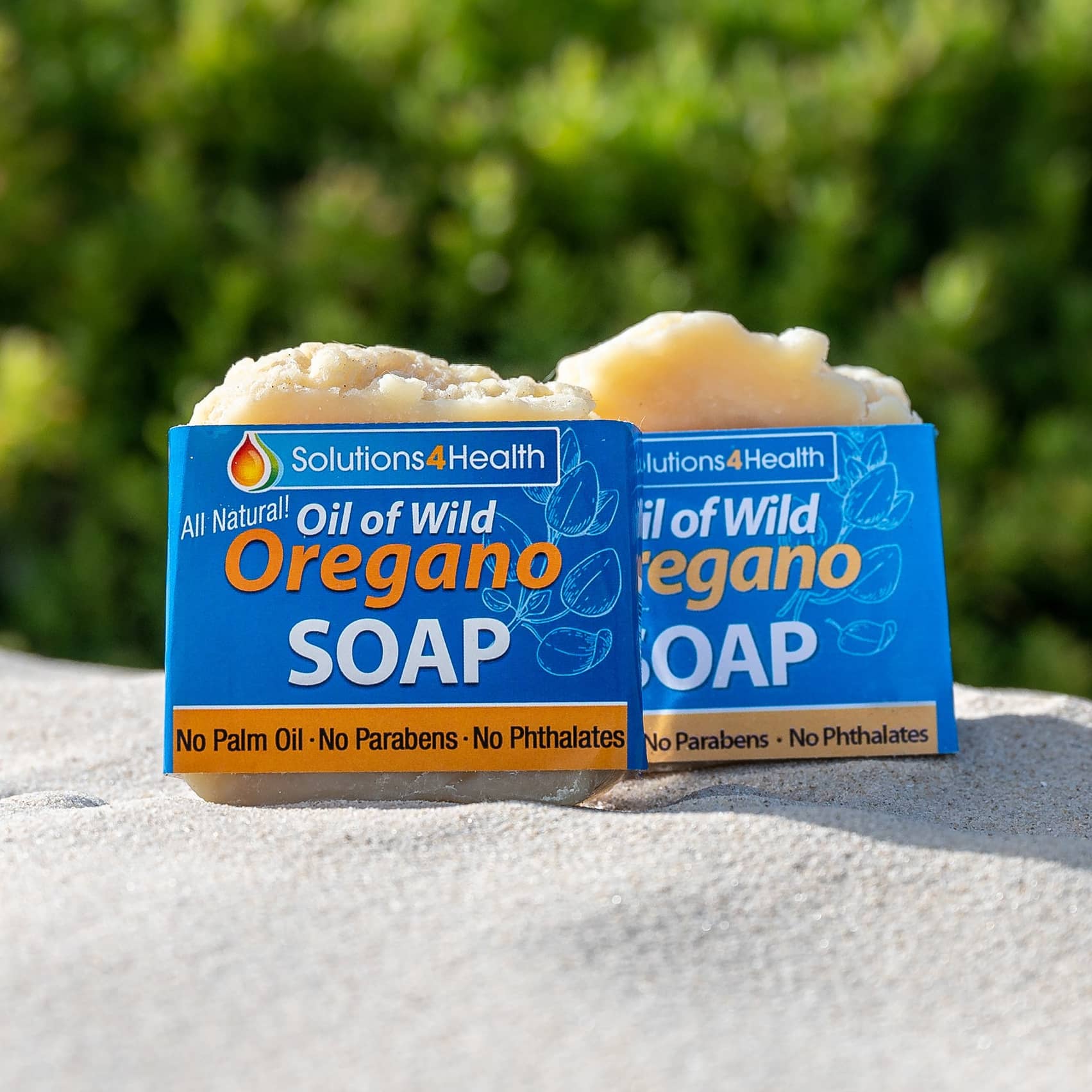 Two bars of Oil of Wild Oregano soap with packaging on a stone surface with greenery in the background.